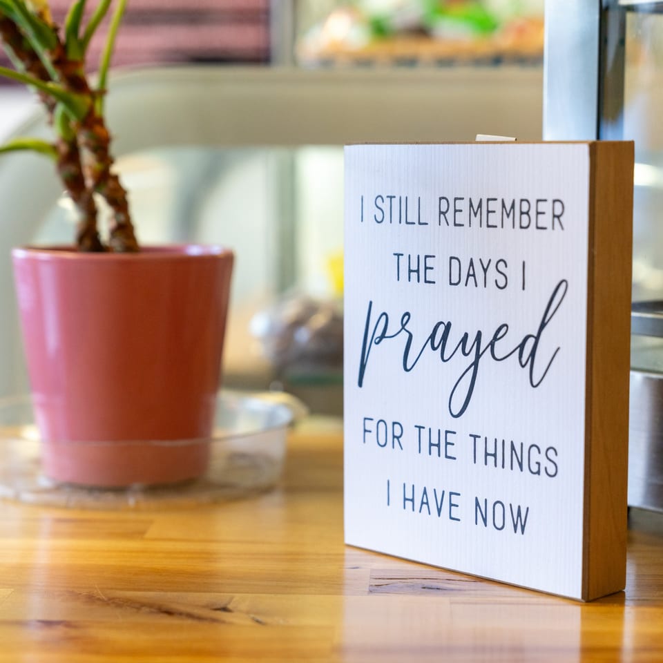 A decorative sign with an inspirational quote sits on a wooden table next to a potted plant.