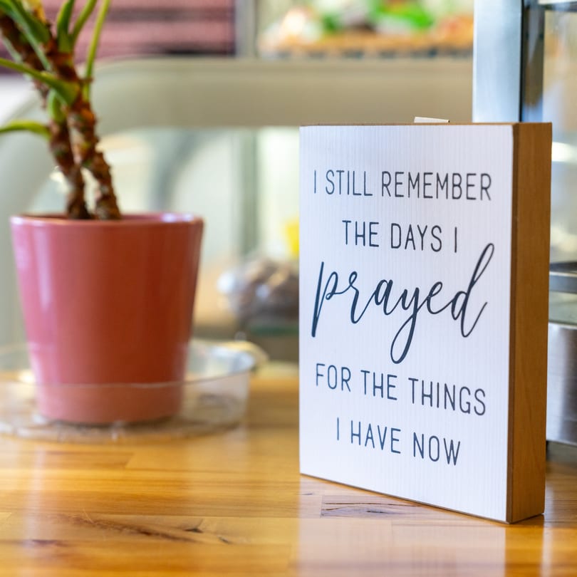 A decorative sign with an inspirational quote sits on a wooden table next to a potted plant.