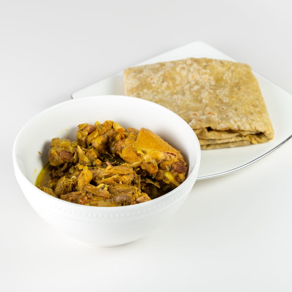 A white bowl of Caribbean curry chicken with a folded roti on a white plate.