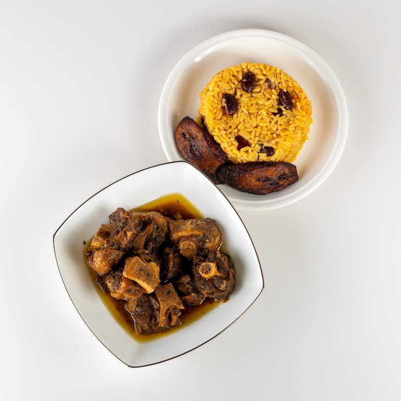Oxtail stew in a white bowl, with yellow rice and plantains in another bowl.
