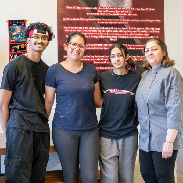 Four smiling Taste of Trini staff members, including a chef, stand together in front of a red wall.