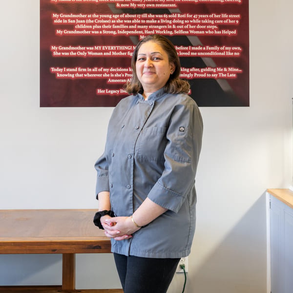 Taste of Trini owner, a woman in a gray chef's coat, stands in front of a red sign.