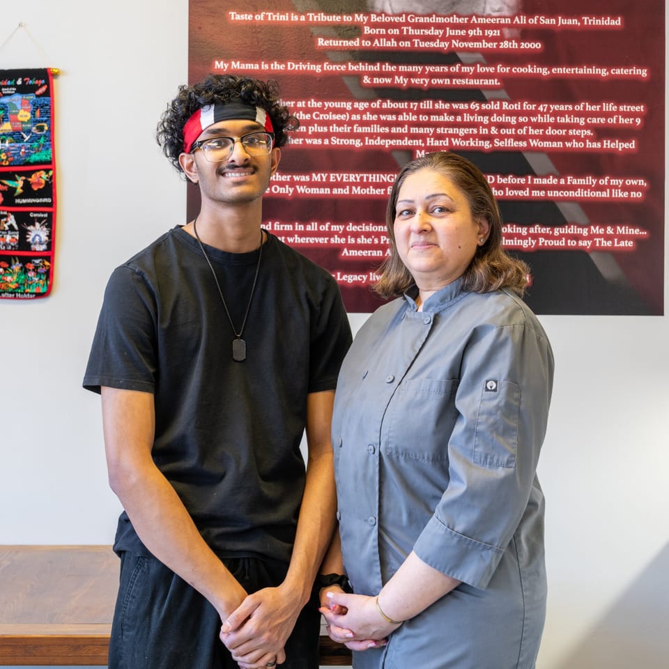 A young man and a woman in a chef's coat stand in front of a tribute wall at Taste of Trini.