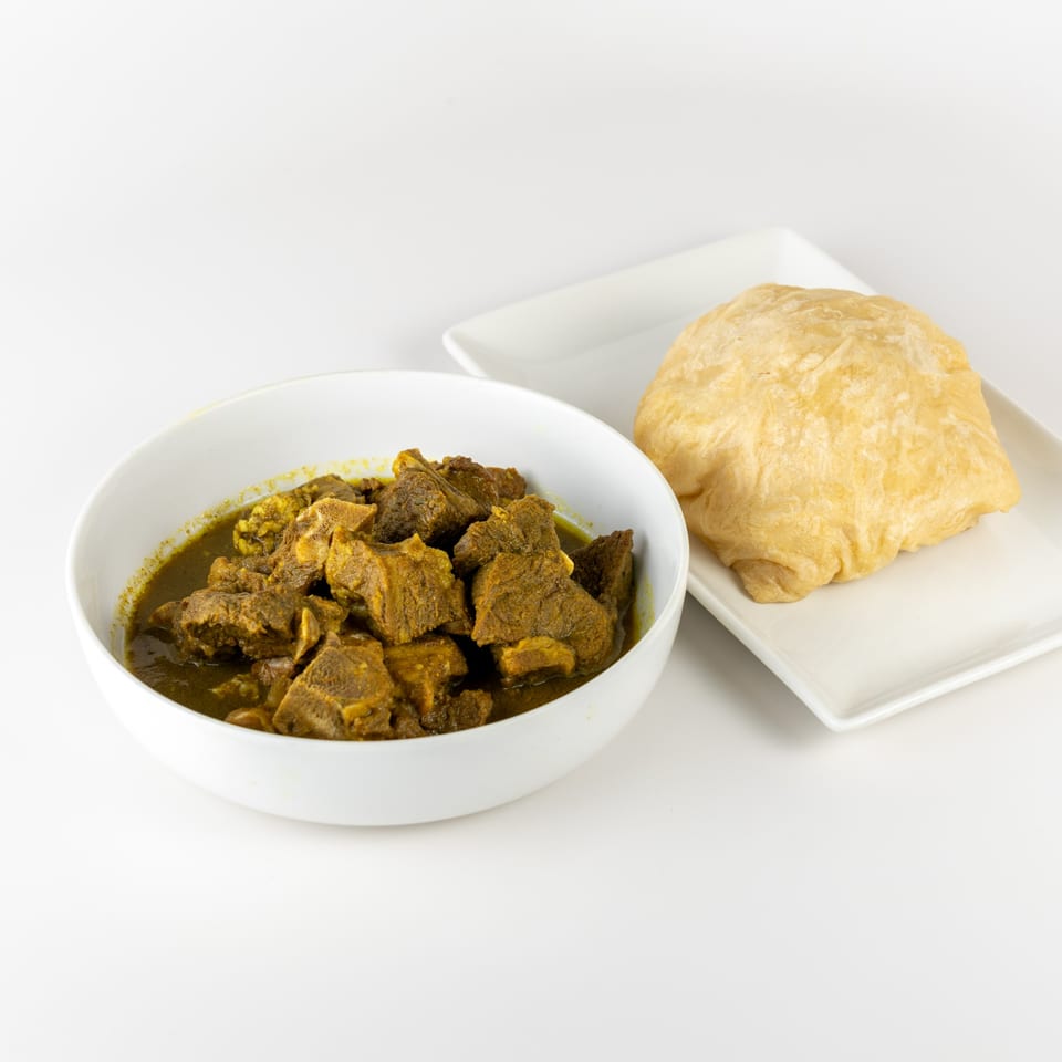 A white bowl of Trini curry goat stew next to a piece of fried bake on a white plate.
